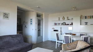 A living room with a grey sofa, a white coffee table, and a floral armchair. There is a dining area with a table and chairs, a kitchen with a fridge, and a bookshelf with books and decorative items. - 2 Person Apartment With sea View (De Koog)