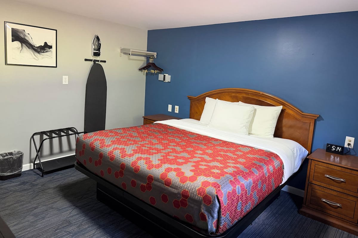 Una habitación de hotel con cama, mesita de noche, cómoda, calentador de pared, reloj de pared y un cuadro enmarcado.