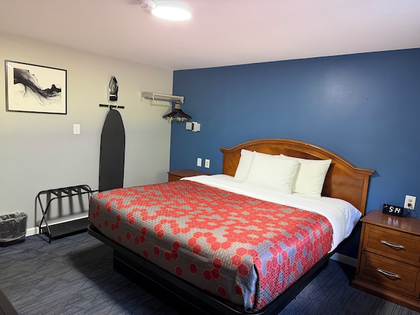 A hotel room with a bed, a nightstand, a dresser, a wall-mounted heater, a wall-mounted clock, and a framed picture on the wall.