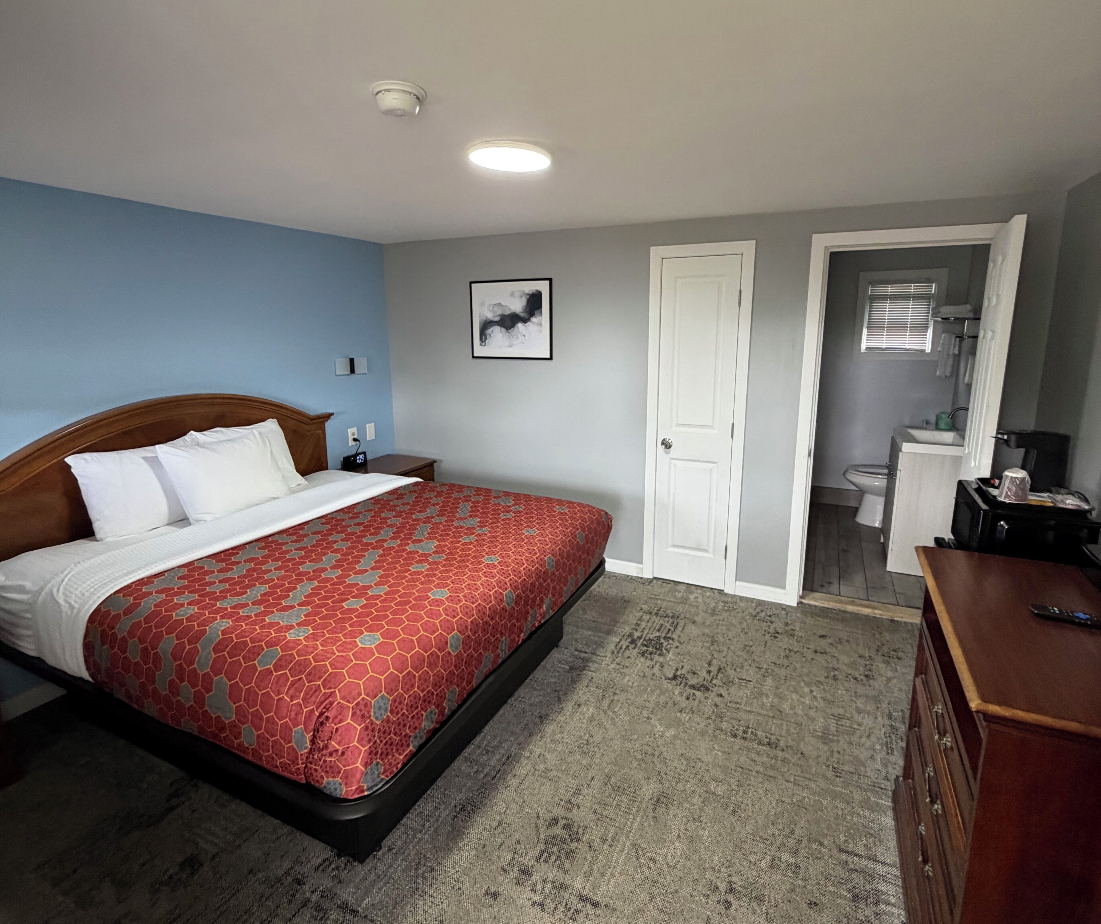 Une chambre à coucher comprenant un lit, une table de chevet, une commode, une porte donnant sur une salle de bain et un luminaire au plafond.