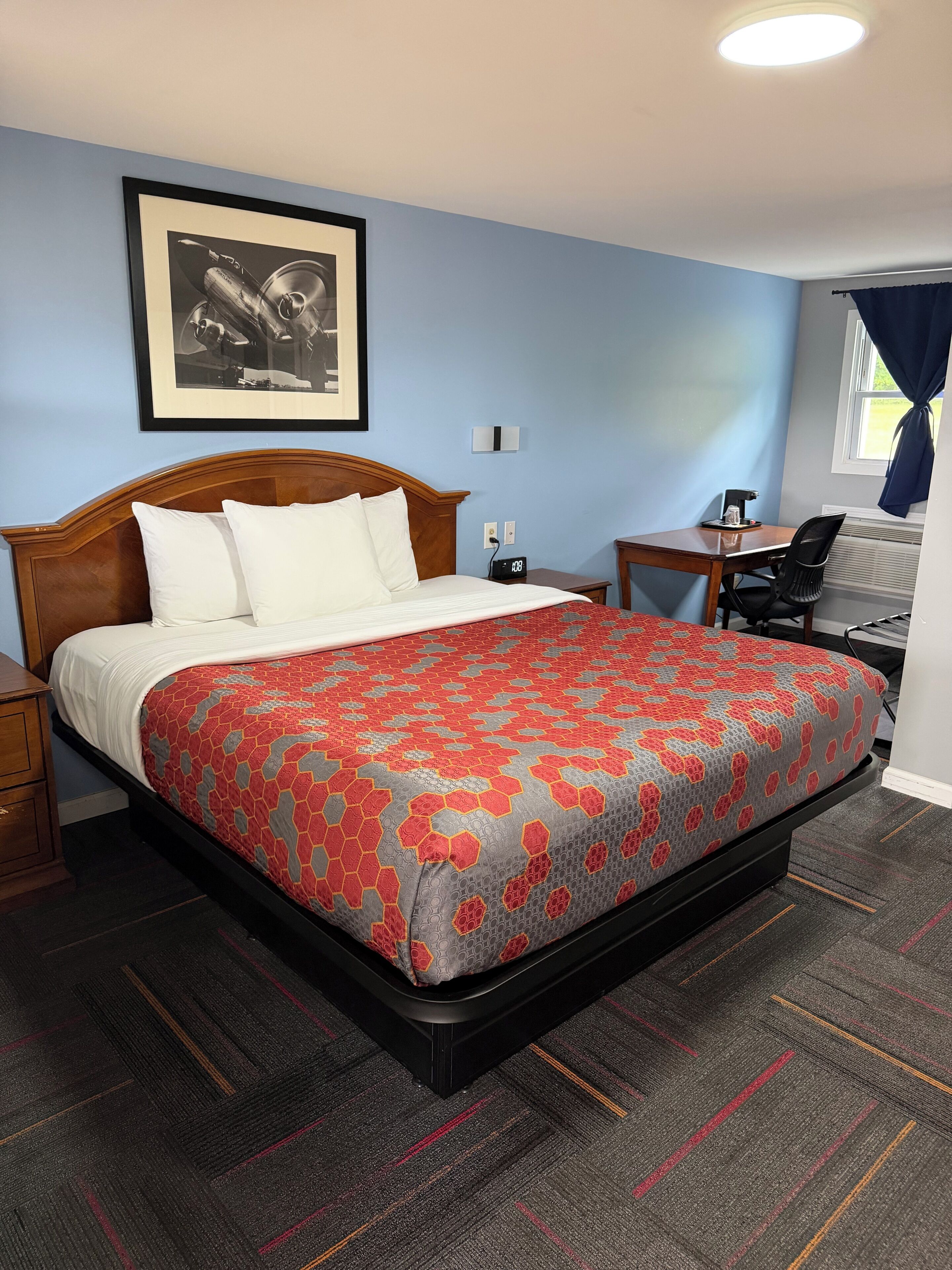 A hotel room with a bed, a desk, a chair, a framed picture on the wall, and a window with curtains.