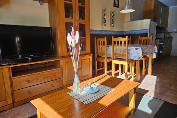 A wooden dining table with a vase of flowers, a television, wooden cabinets, and a kitchen area.
