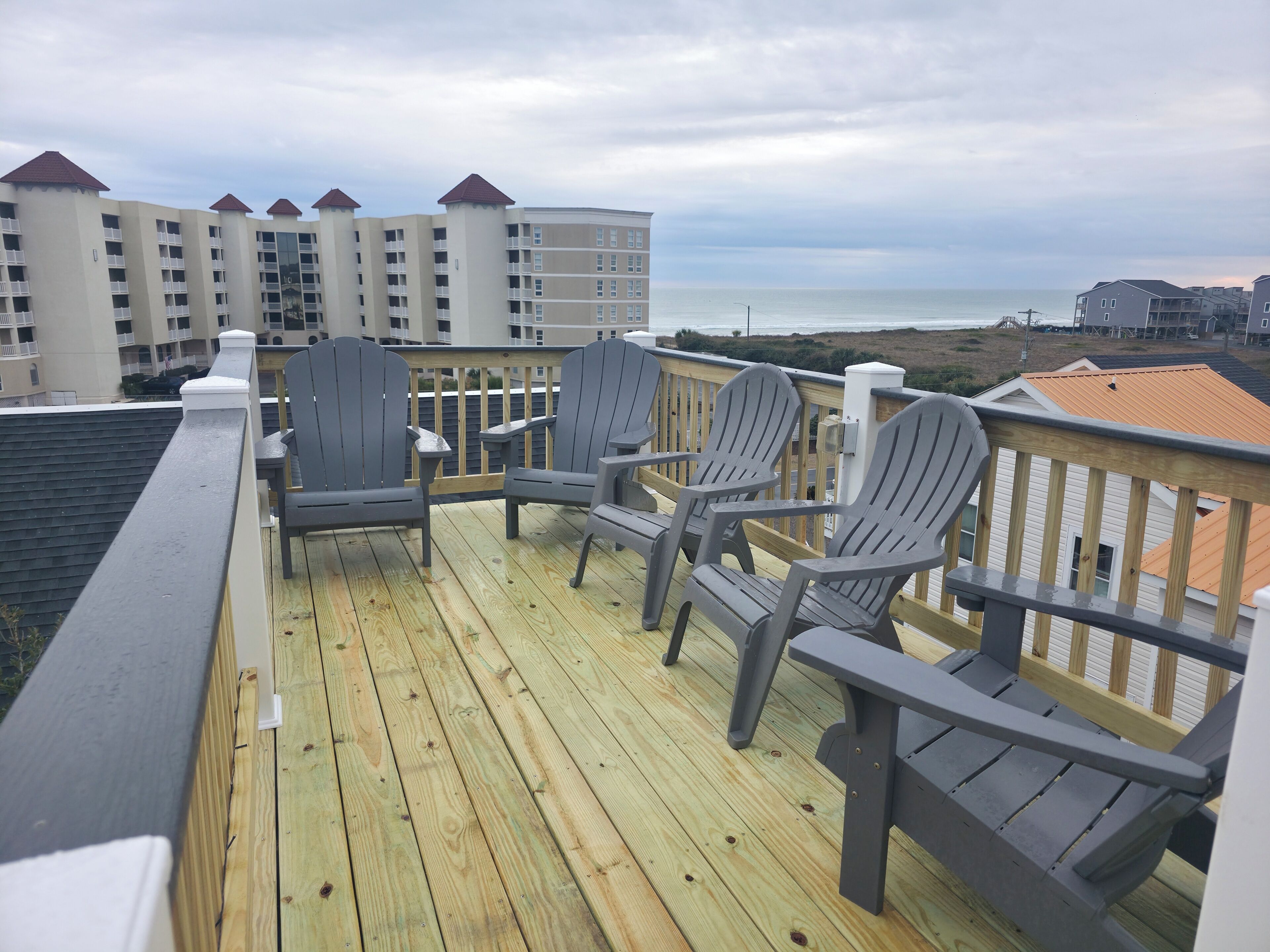 A wooden deck with Adirondack chairs overlooking a beach and buildings.