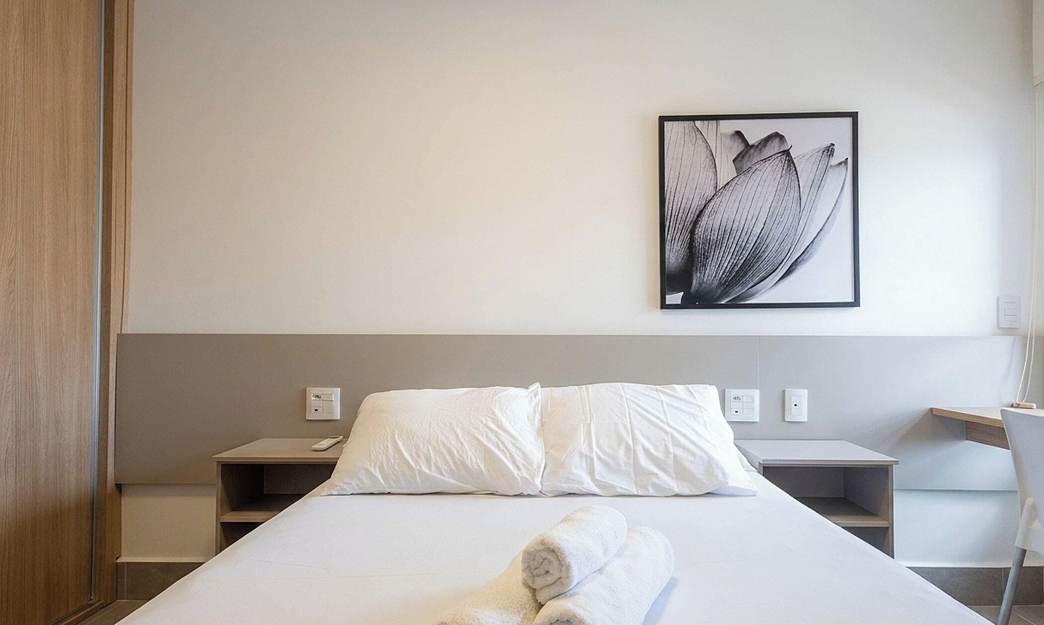 Habitación de hotel con cama, ropa de cama blanca, un cuadro colgado en la pared, mesitas de noche y un escritorio con silla.