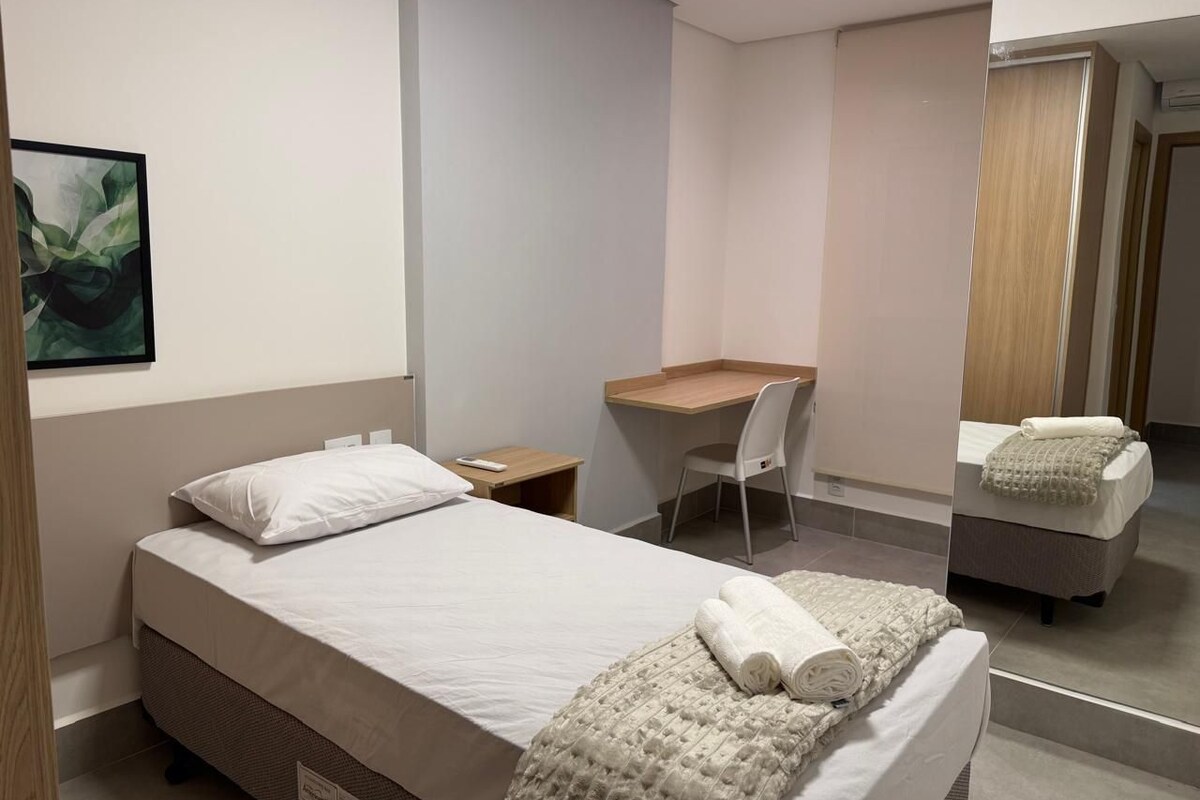 A modern hotel room with a bed, a desk, a chair, a mirror, and a bathroom.