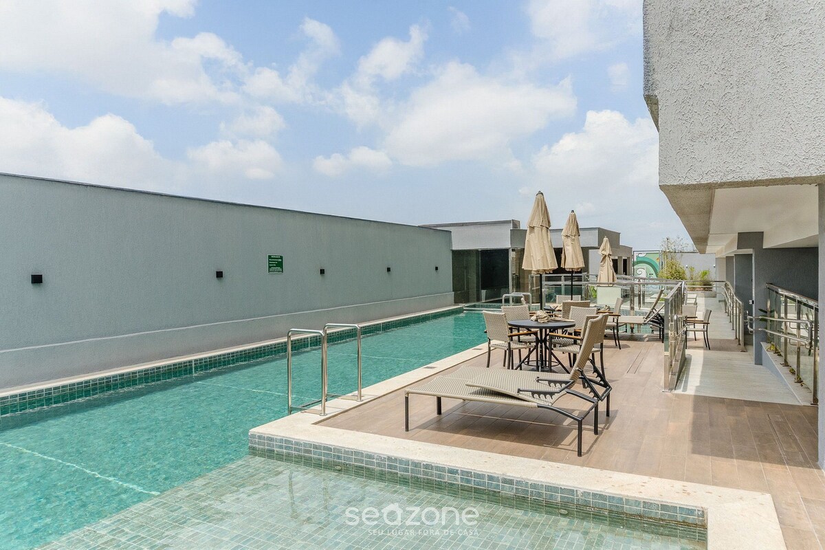 A rooftop pool area with lounge chairs and umbrellas, surrounded by a white building.