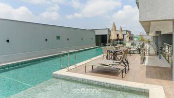A rooftop pool area with lounge chairs and umbrellas, surrounded by a white building.