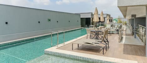 A rooftop pool area with lounge chairs and umbrellas, surrounded by a white building.