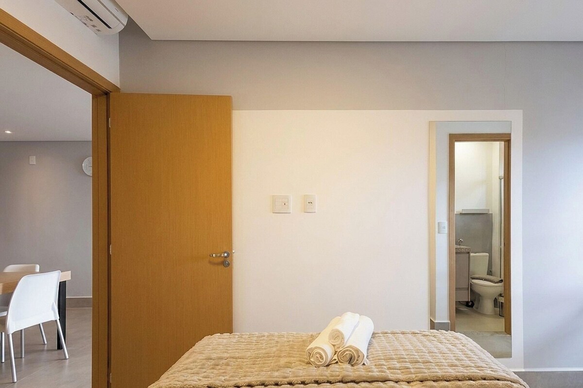 A hotel room with a bed, a wooden door, a white wall, and a bathroom visible through an open door.