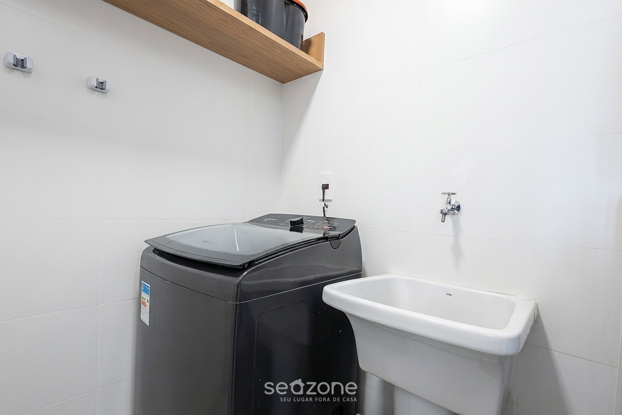 A compact bathroom with a black washing machine, a white sink, and a wooden shelf above the machine.