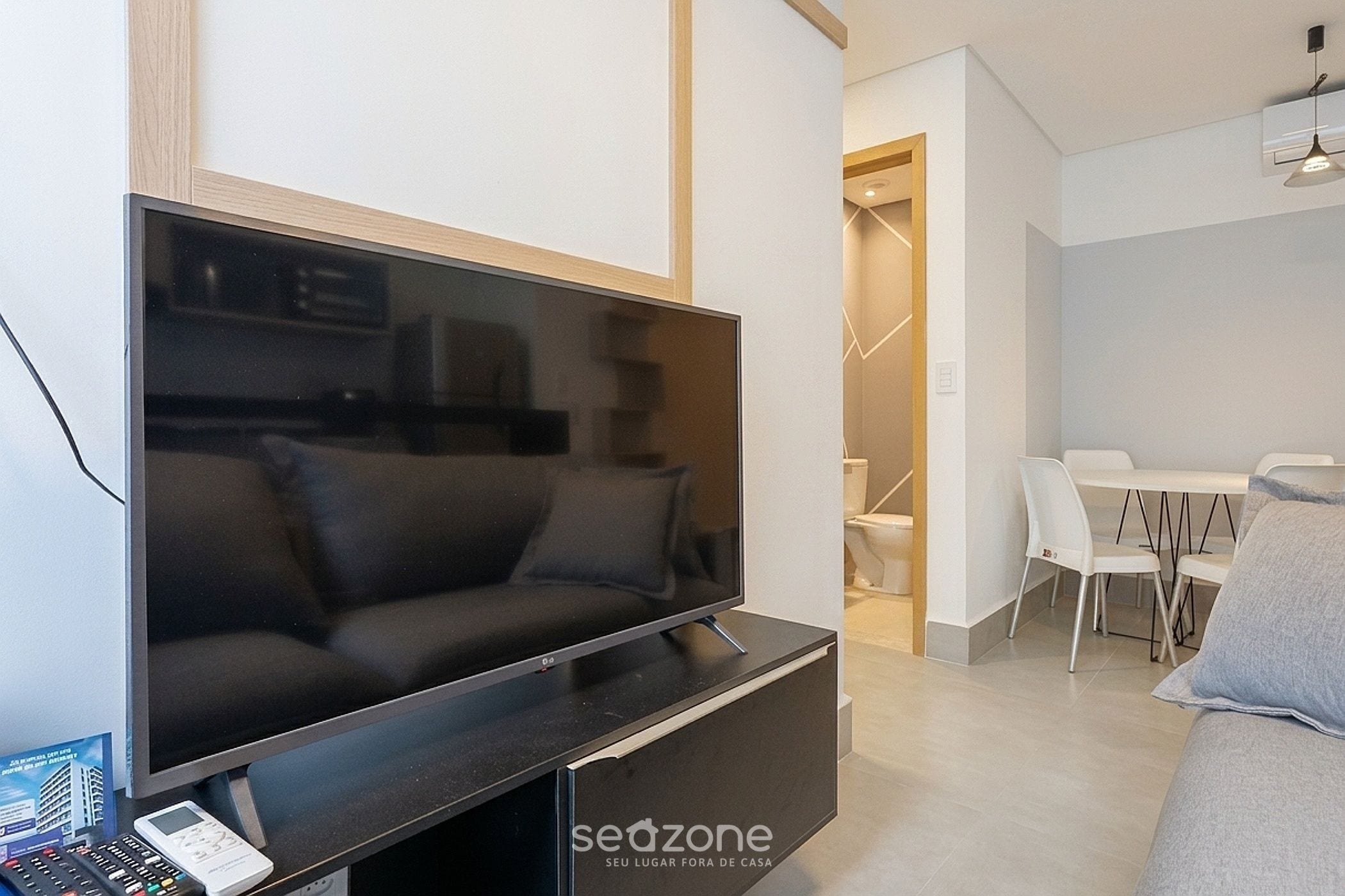 A modern living room with a large flat-screen TV, a sofa, and a dining area with white chairs and a table.