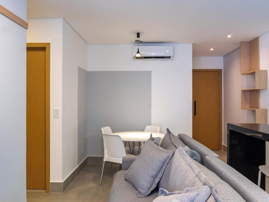A modern living room with a sofa, a dining table with chairs, a kitchen area with a counter and cabinets, and an air conditioning unit on the ceiling.