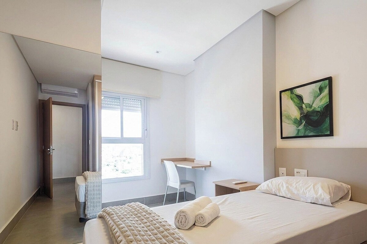 A modern hotel room with a bed, a desk, a chair, and a painting on the wall.