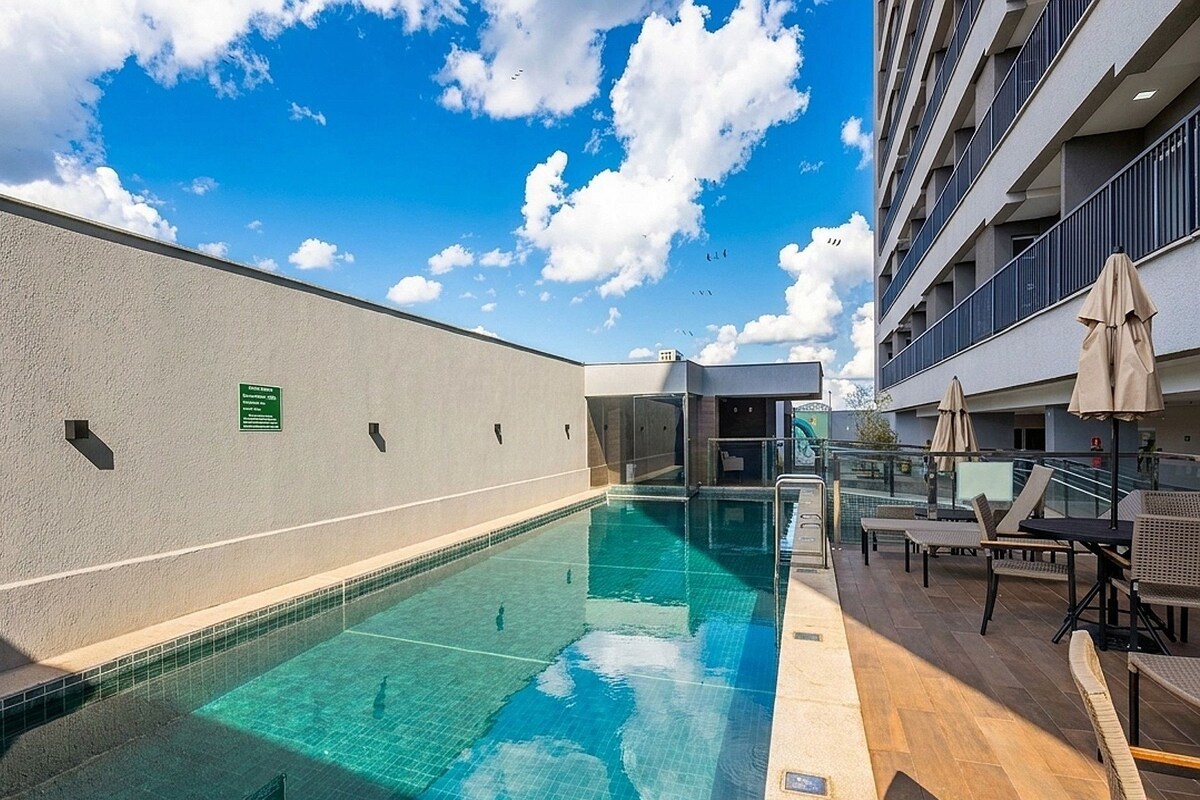A swimming pool with clear blue water, surrounded by a low wall and a building with balconies.