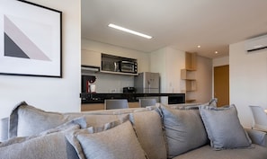 A modern living room with a grey sofa, a kitchen in the background, and a framed picture on the wall.