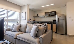 A modern living room with a sofa, a flat-screen TV, and a kitchen area with a refrigerator, microwave, and coffee machine. - Apts in a Building With Pool and Gym - GRL (Anapolis)