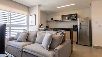 Una sala de estar moderna con sofá, televisor de pantalla plana y área de cocina con refrigerador, microondas y cafetera.