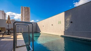 Une piscine avec un escalier en métal, un espace de détente avec des chaises et un parasol, et un immeuble de grande hauteur en arrière-plan.
