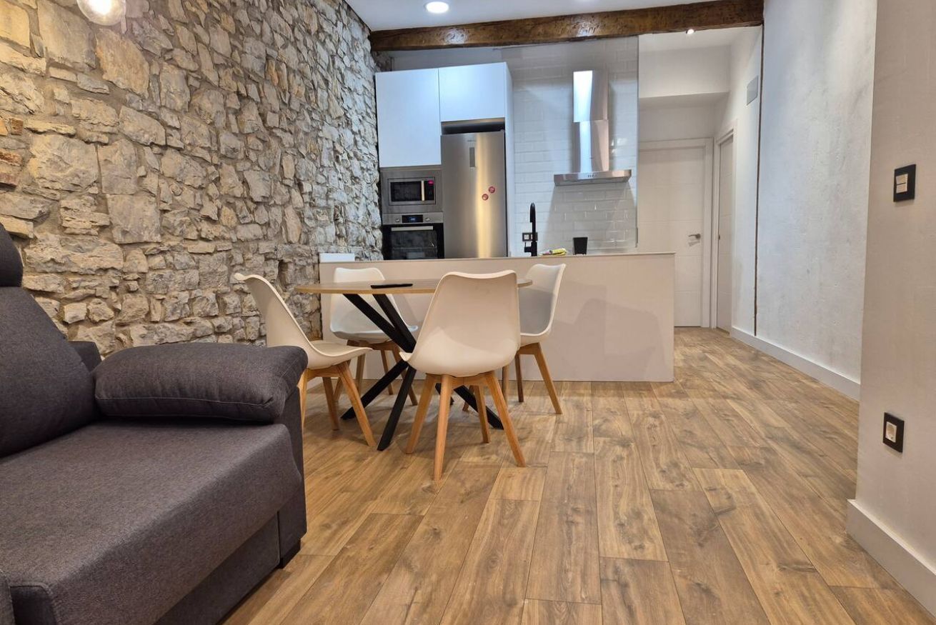 Una sala de estar moderna con una pared de piedra, un sofá gris, una mesa de comedor de madera con sillas y una cocina al fondo.