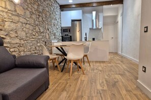 Un salon moderne avec un mur en pierre, un canapé gris, une table à manger en bois avec des chaises, et une cuisine en arrière-plan.