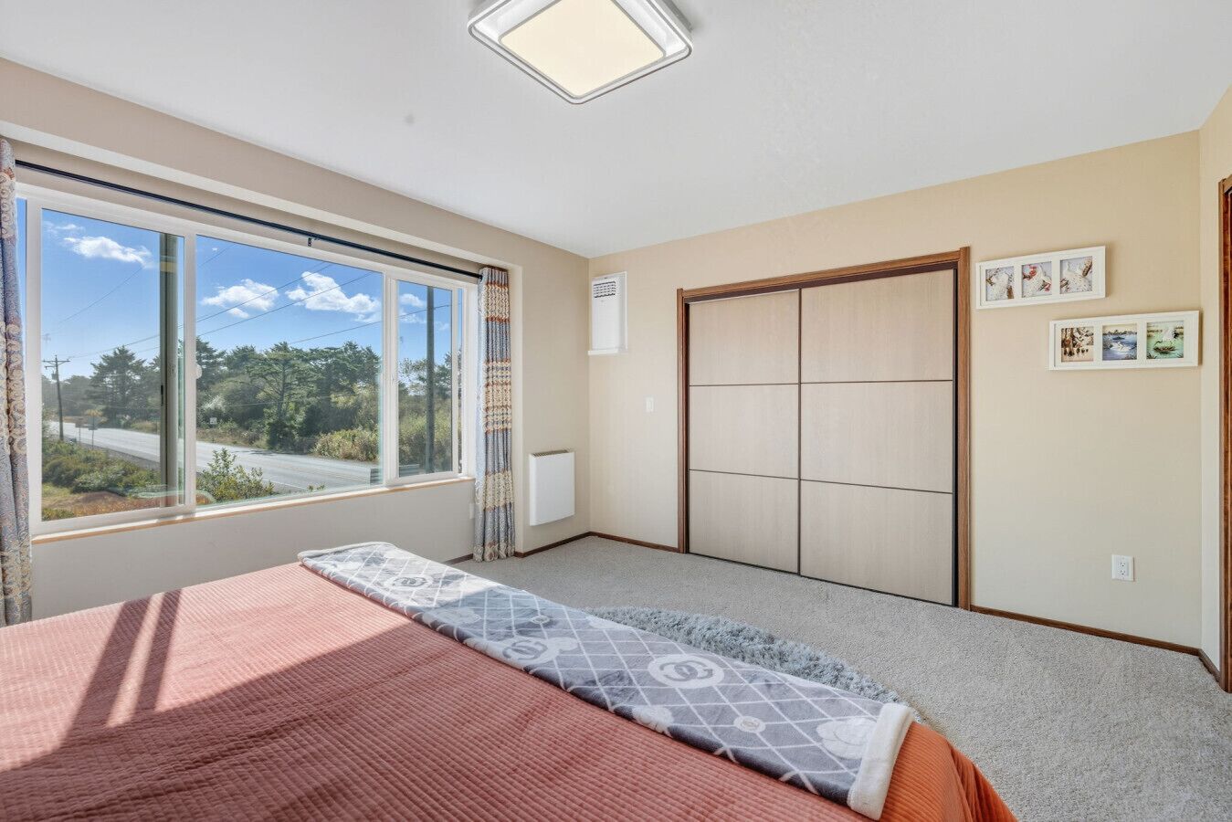 Une chambre avec une grande fenêtre, un lit recouvert d’une couvre-lit à motifs, un radiateur et un placard à portes coulissantes.