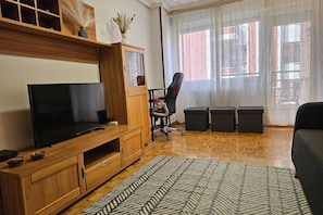 A living room with a TV, wooden cabinet, office chair, ottomans, and a patterned rug.