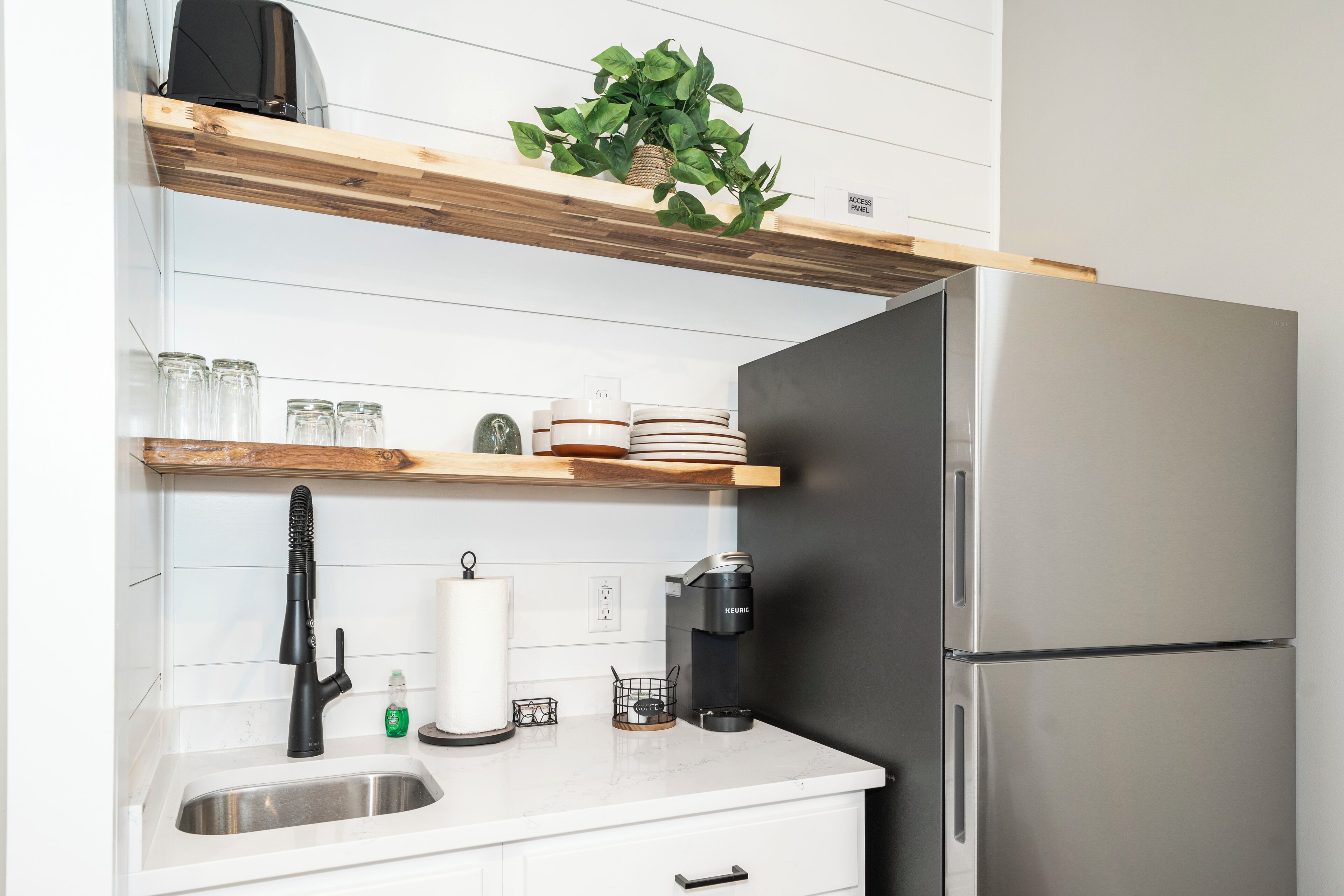 Una cocina con un refrigerador de acero inoxidable, una cafetera, un lavamanos y estantes de madera con frascos y platos de vidrio.