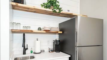 Una cocina con un refrigerador de acero inoxidable, una cafetera, un lavamanos y estantes de madera con frascos y platos de vidrio.