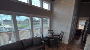 A spacious living room with a dark leather sofa, a wooden coffee table, and a dining area with a table and chairs. Large windows provide natural light and views of the outdoors. - Carolina Camp Cedar (Pickens)