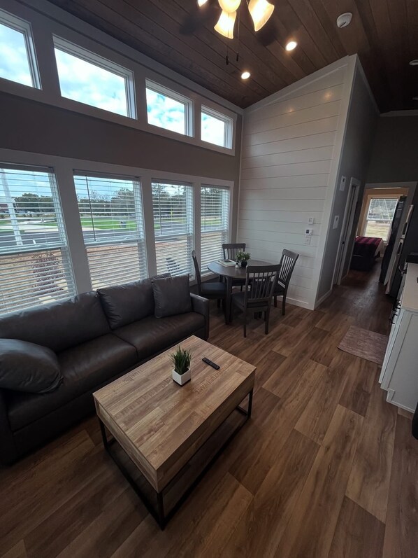 A spacious living room with a dark leather sofa, a wooden coffee table, and a dining area with a table and chairs. Large windows provide natural light and views of the outdoors. - Carolina Camp Cedar (Pickens)