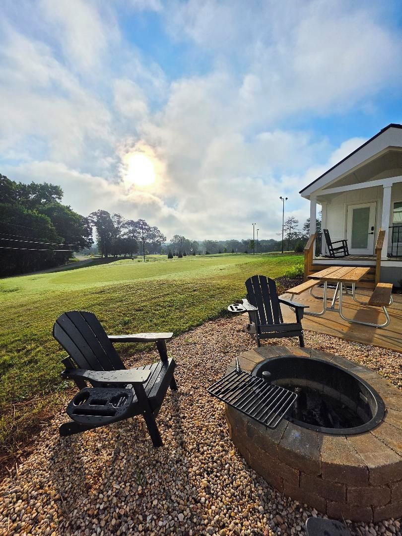 An outdoor seating area with Adirondack chairs and a fire pit, overlooking a grassy field and a building with a covered patio.