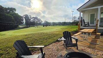 An outdoor seating area with Adirondack chairs and a fire pit, overlooking a grassy field and a building with a covered patio.