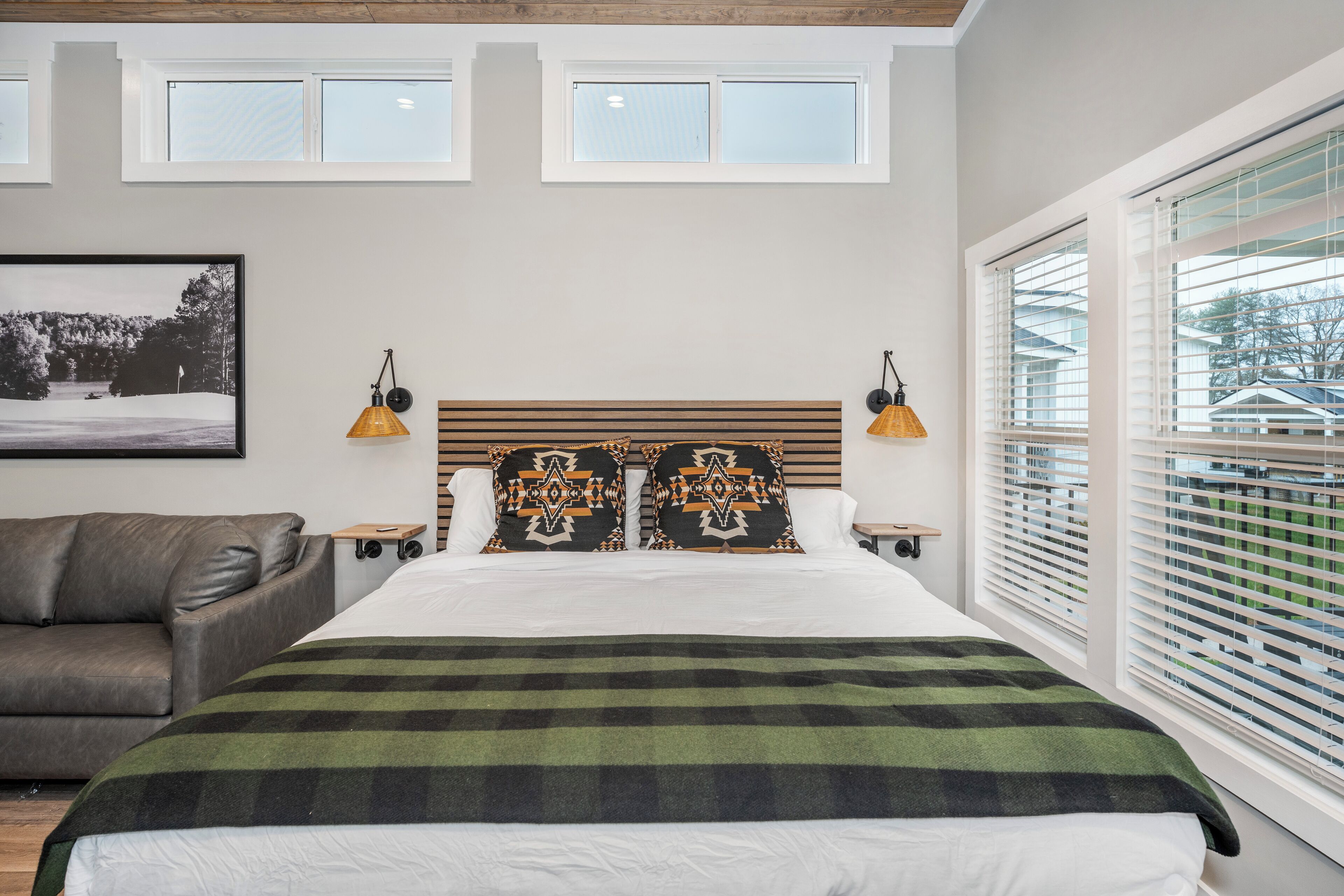 Een slaapkamer met een groot bed, twee nachtkastjes, een houten hoofdbord en een raam met luxaflex.