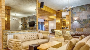 Un salón amplio con un sofá beige grande, una mesa de centro de madera y una chimenea de piedra. Ofrece múltiples opciones de asientos, incluyendo un sillón con cojín y un sofá de cuero. La habitación presenta vigas de madera a la vista y paredes de piedra, creando un ambiente rústico.