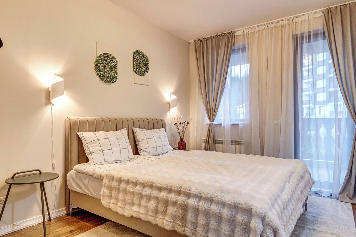 Un dormitorio con cama, lámparas de noche, una mesita y una ventana con cortinas.