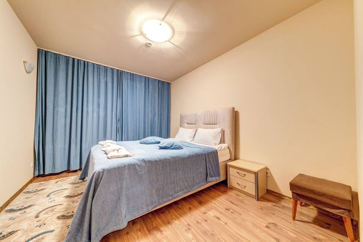 Una cama bien tendida con una colcha azul, una mesita de noche con una lámpara, y un banco junto a la ventana.