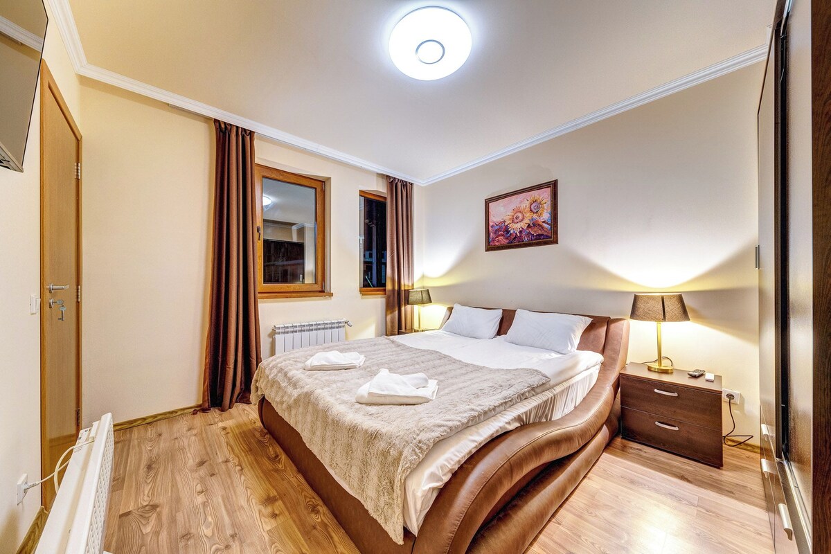 Een hotelkamer met een groot bed, een nachtkastje met een lamp, een raam met gordijnen, een radiator en een schilderij aan de muur.