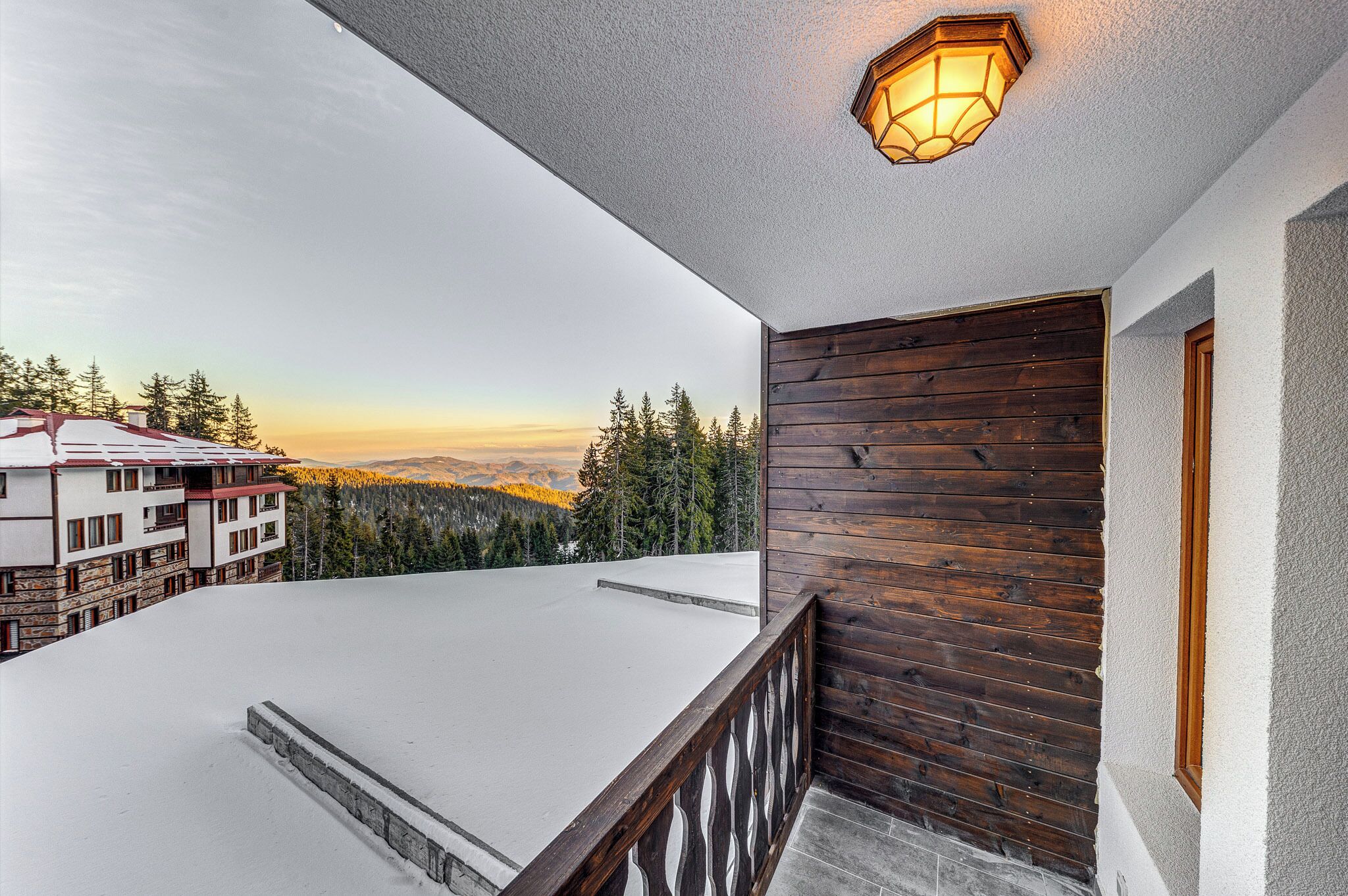 A balcony with a wooden railing, a snow-covered roof, and a view of a forest and buildings in the distance.