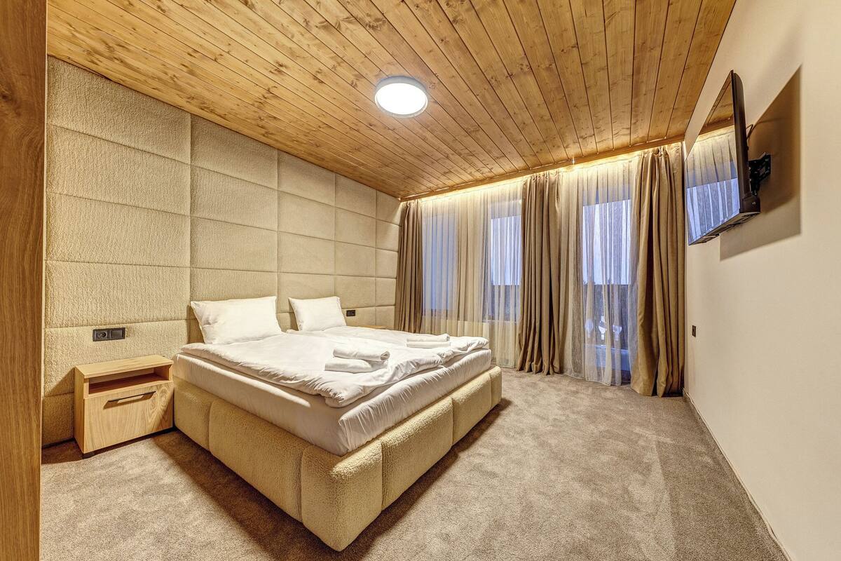 Un dormitorio con techo de madera, una cama, una mesita de noche, un televisor y una ventana con cortinas.
