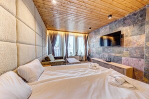 Habitación de hotel con una cama grande, un televisor de pantalla plana montado en una pared de piedra, techo de madera y una ventana con cortinas.