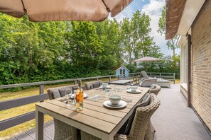 An outdoor patio with a wooden table set for dining, wicker chairs, and a thatched umbrella. - Charming Country House for 4 People (De Koog)