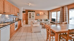 A spacious kitchen with wooden cabinets, a dining area with a wooden table and chairs, and a living room with a sofa and a television. - Pistes du Soleil C1 - Four Bedroom Apartment, Sleeps 8 (Le Grand Bornand)