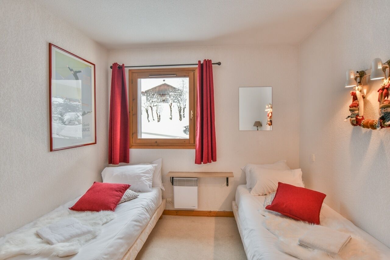 Ein Schlafzimmer mit zwei Betten, einem Fenster mit roten Vorhängen, einem an der Wand hängenden Bild, einem Nachttisch mit einer Lampe und einem Wandspiegel.
