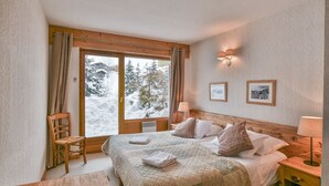 A cozy bedroom with a large bed, a wooden chair, a nightstand, and a window with curtains. - Pistes du Soleil C1 - Four Bedroom Apartment, Sleeps 8 (Le Grand Bornand)