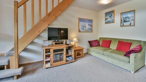Un salon avec un escalier en bois, un canapé vert agrémenté de coussins rouges, un téléviseur à écran plat, un meuble de rangement en bois et des tableaux encadrés accrochés au mur.