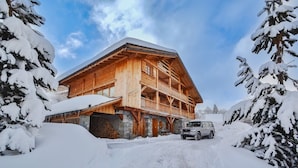 A large wooden chalet with a stone foundation, a balcony, and a snow-covered driveway. - Chalet Mitoyen - Solar Est (Le Grand Bornand)