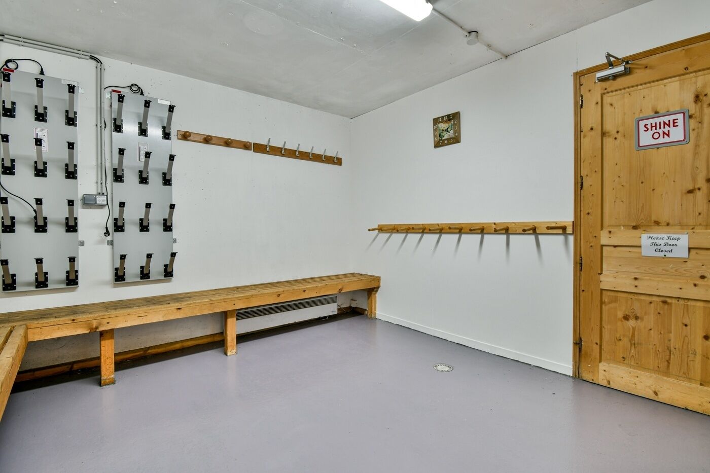 Ein Zimmer mit einer hölzernen Sitzbank, Kleiderhaken, einer Uhr und einer Tür mit der Aufschrift 'SHINE ON'.