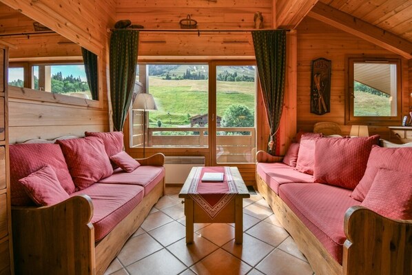 A cozy wooden cabin with two red sofas, a small table, and large windows offering a view of a green landscape. - Pistes du Soleil A15 (Le Grand Bornand)