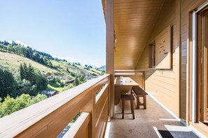 A wooden balcony with a table and chairs, overlooking a valley with houses and trees.