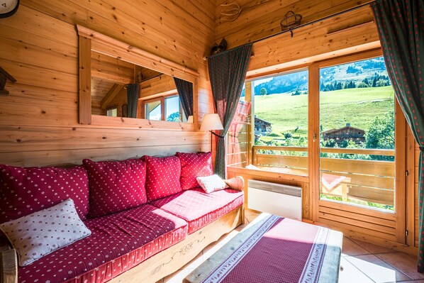 Interior de una cabaña de madera con un sofá, una ventana que ofrece vistas a un paisaje verde y una lámpara.